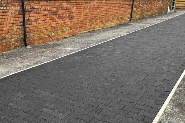 A wide outdoor path made of black rubber pavers with a brown brick wall behind it