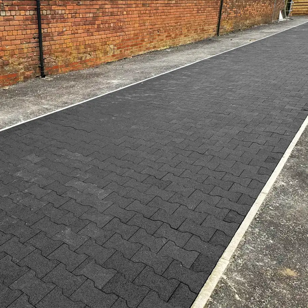 A wide outdoor path made of black rubber pavers with a brown brick wall behind it