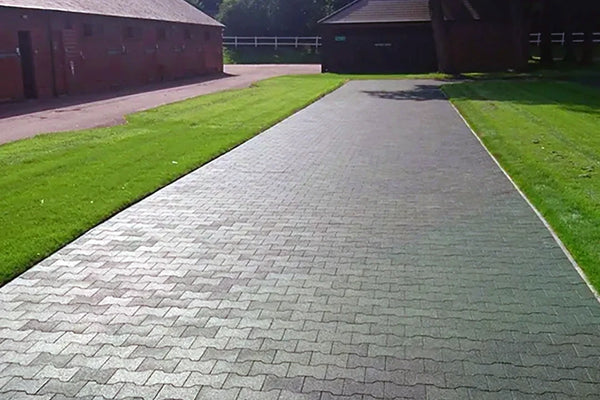 A long wide path made of green rubber pavers outside with some grass and buildings around