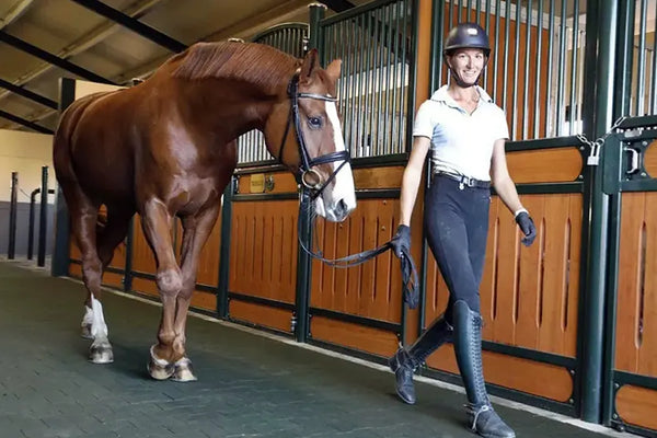 A woman smiling and walking with a horse in a barn on a black rubber paver floor