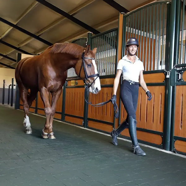 A woman smiling and walking with a horse in a barn on a black rubber paver floor