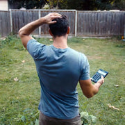 Man in a blue shirt holding a calculator outdoors while scratching his head