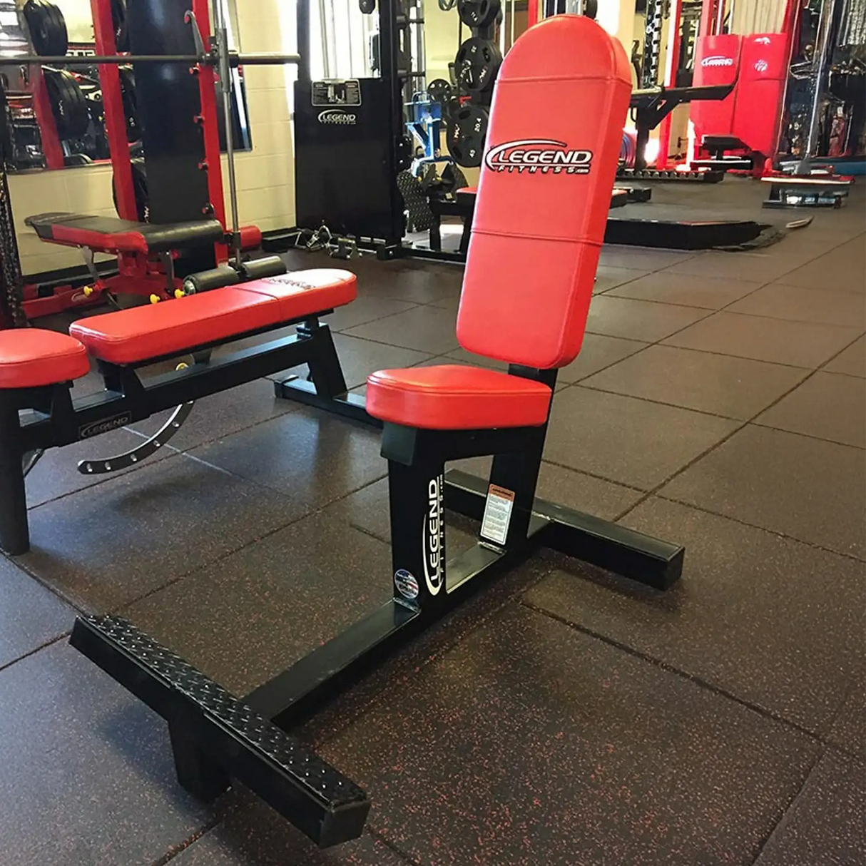 The Legend Fitness 3104 Multi-Purpose Bench in red and black sits on a rubber floor in a spacious gym, with various fitness equipment visible in the background.