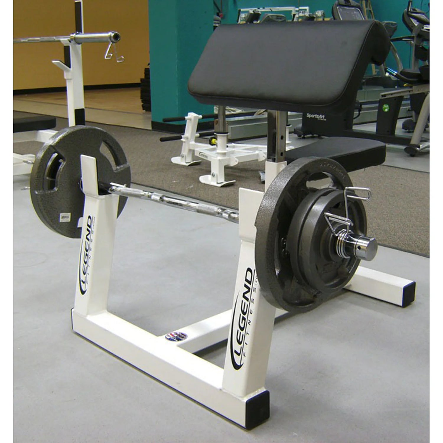 The Legend Fitness 3114 Preacher Curl is shown in a gym, featuring a padded bench and weight rack labeled Legend Fitness, with additional gym equipment visible in the background.