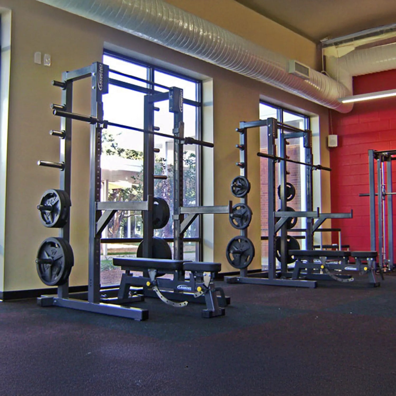 Gym equipment including weight racks and benches in a well-lit room with large windows.