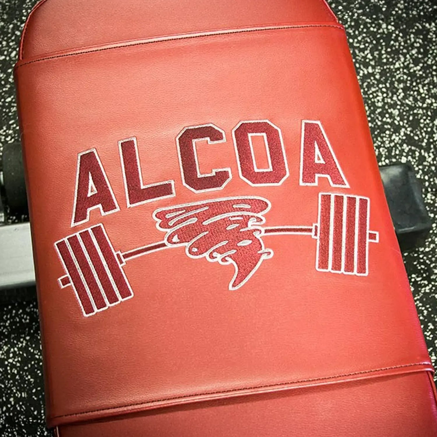 Red weightlifting bench with 'ALCOA' logo featuring a tornado and barbell embroidered on it on a black with white speckled gym floor background.