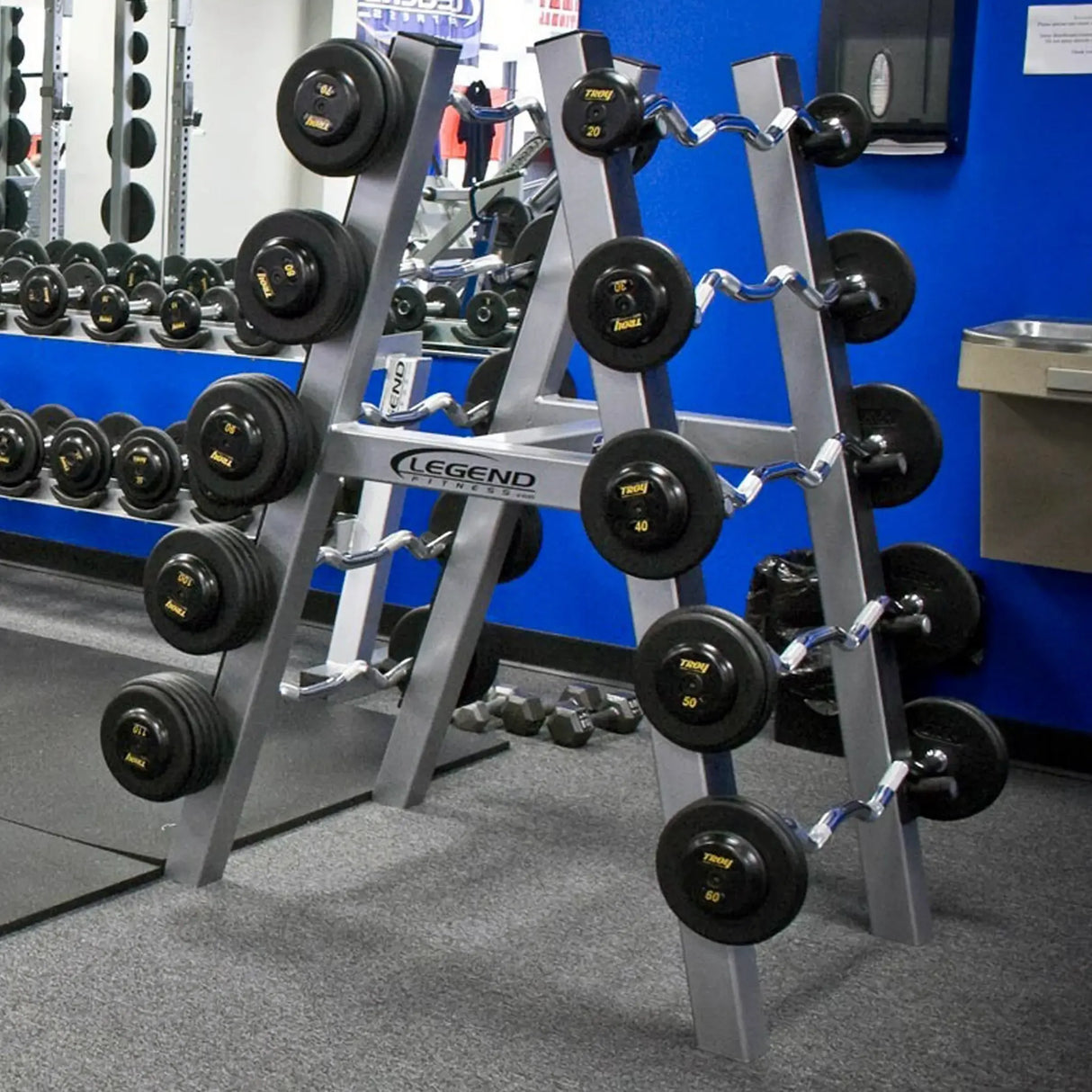 A silver barbell storage rack with ten barbells stored on it in a gym setting with a blue wall in the background