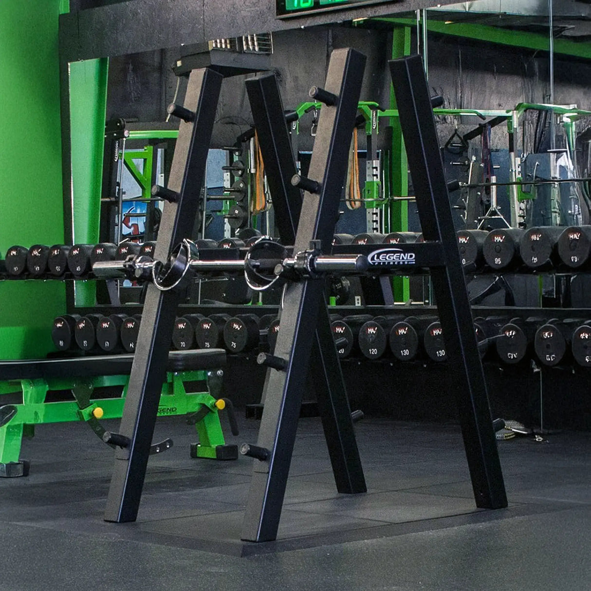 A black barbell storage rack with a barbell stored on it in a gym setting with dumbbells and mirrors in the background