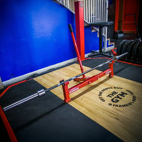 A red metal barbell jack with a silver barbell on it on a wooden platform In a gym setting