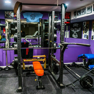Gym interior with weightlifting equipment, including a squat rack and barbell, against a purple wall.