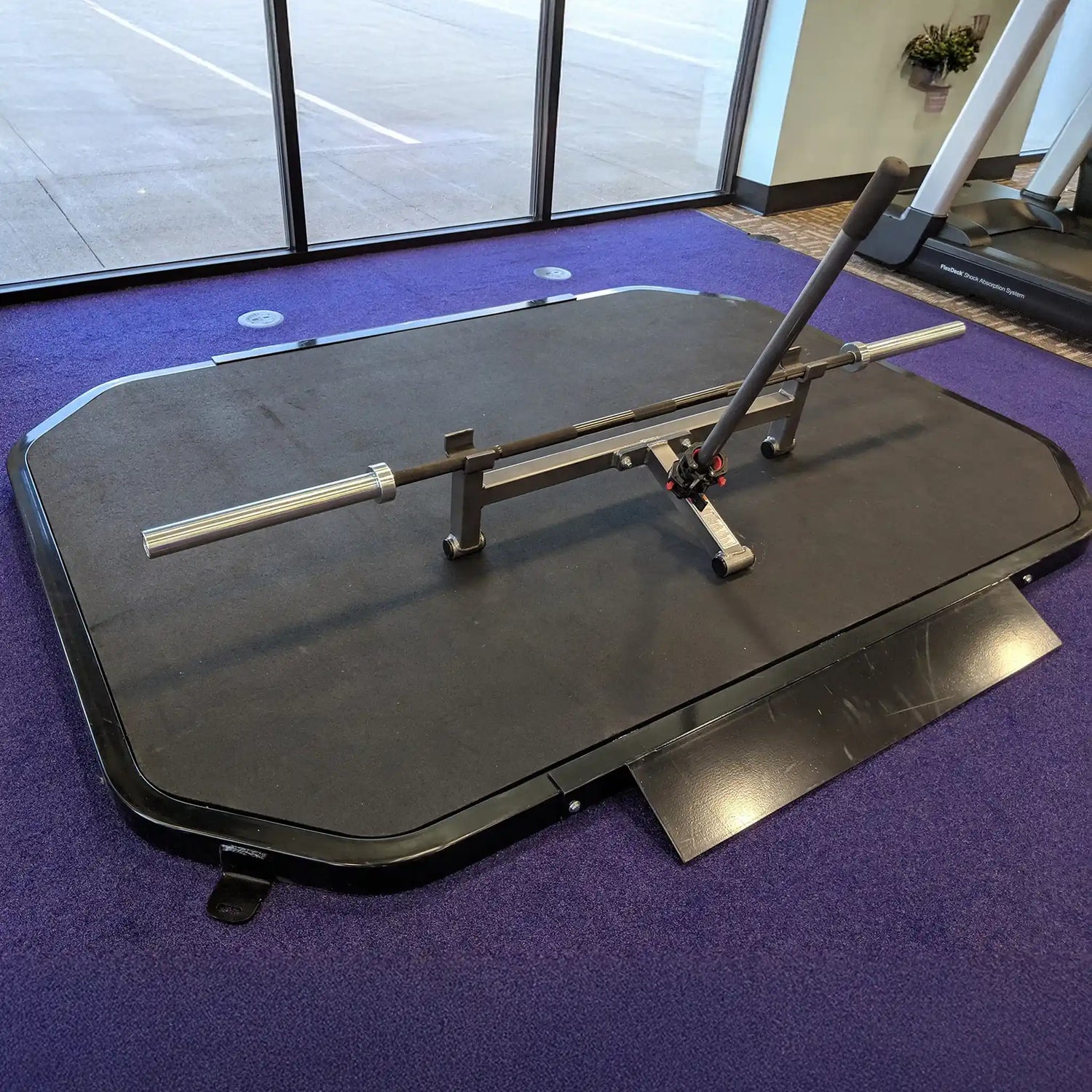 Black weightlifting platform with barbell and a bar jack on a purple floor in a gym setting