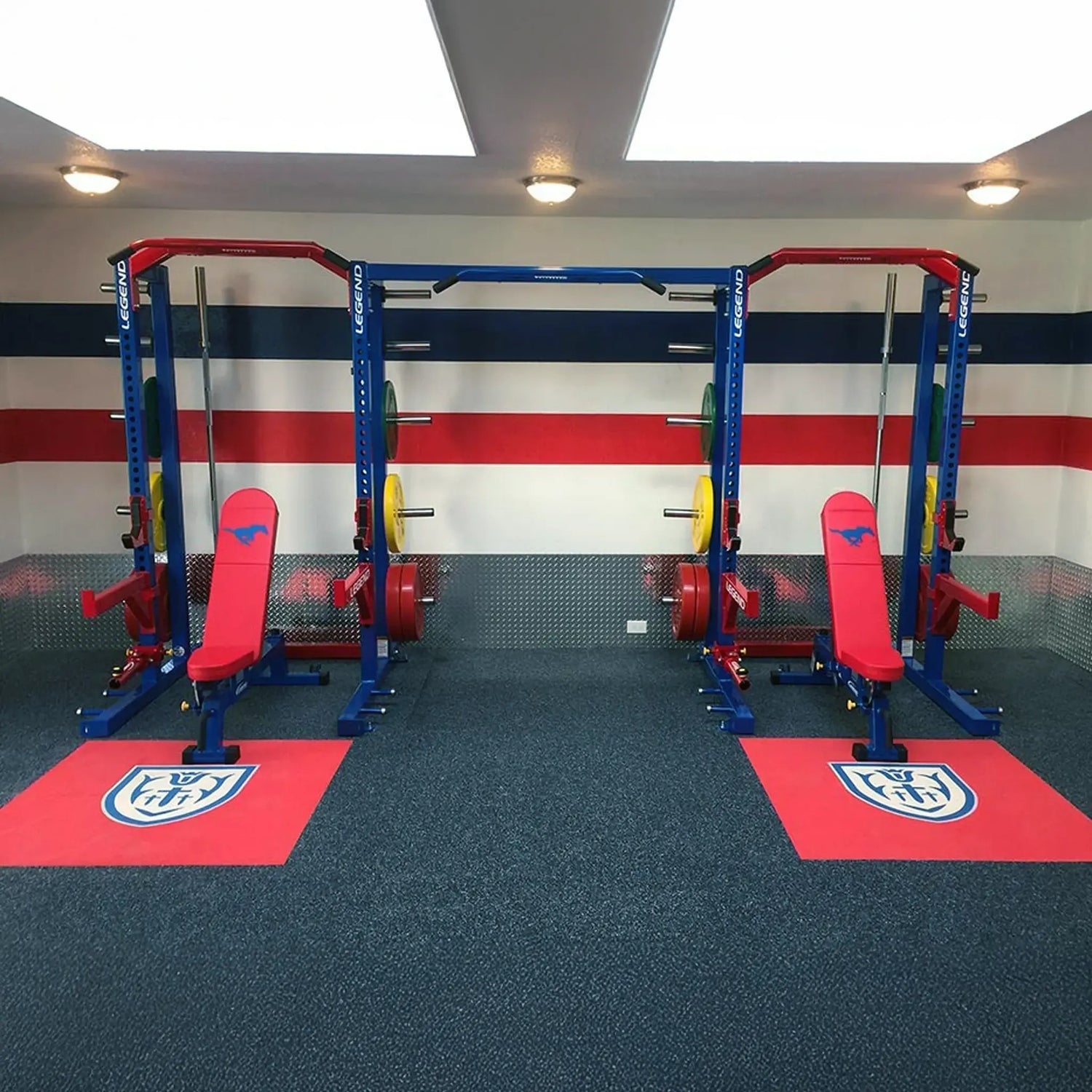 Gym setup with weightlifting racks and benches on red mats against a wall with horizontal stripes.