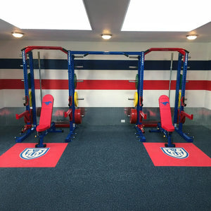 Gym setup with weightlifting racks and benches on red mats against a wall with horizontal stripes.