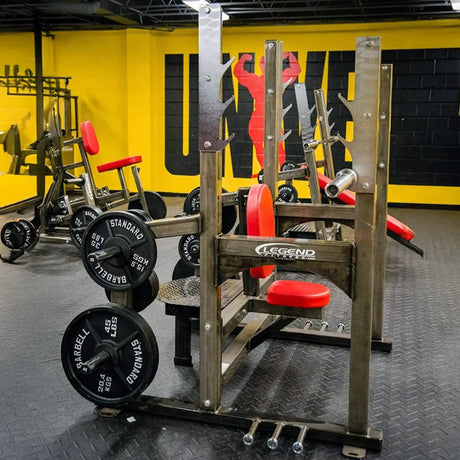An industrial shoulder press weightlifting bench with red pads in a gym setting.