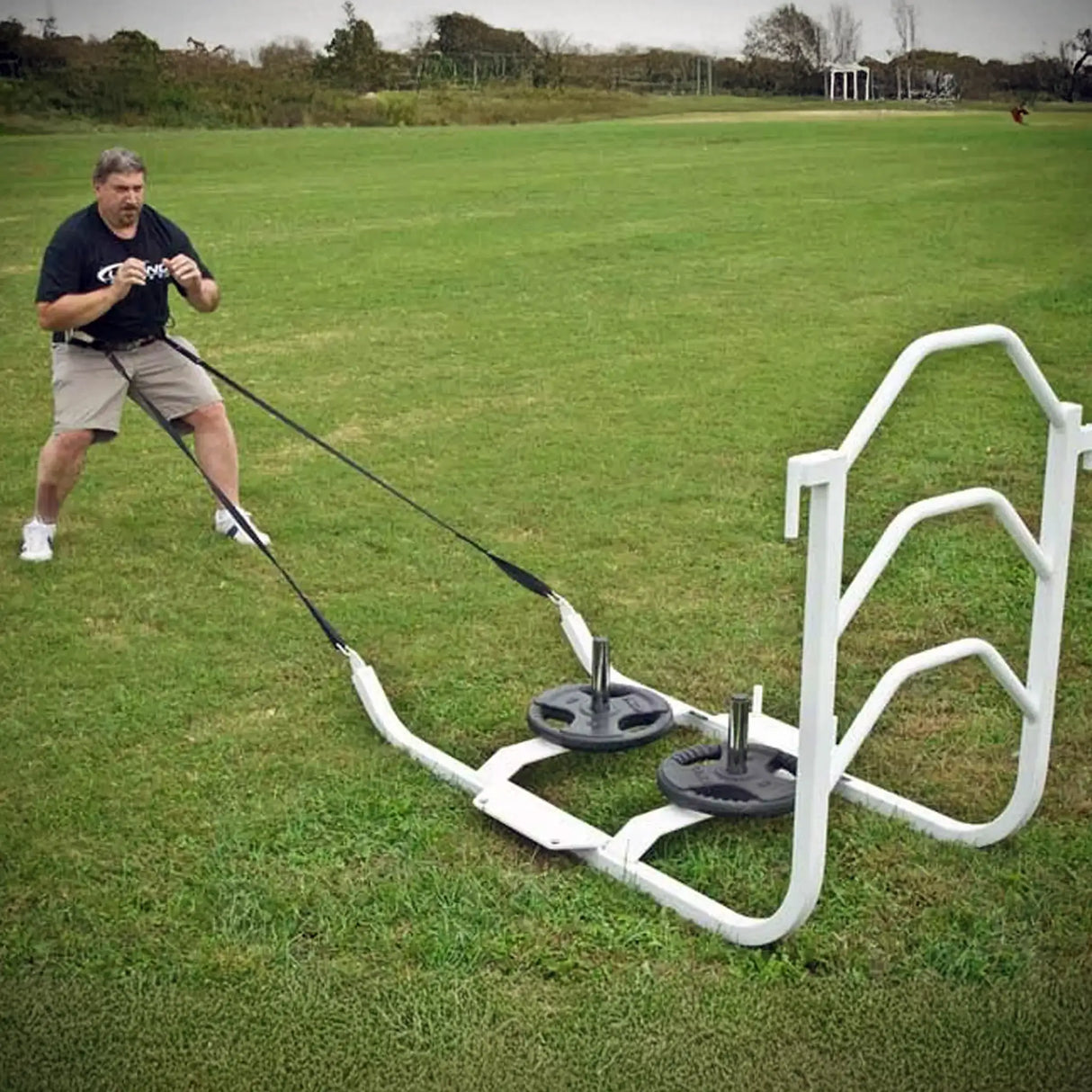 A man pulling a white metal sled on grass by two black straps