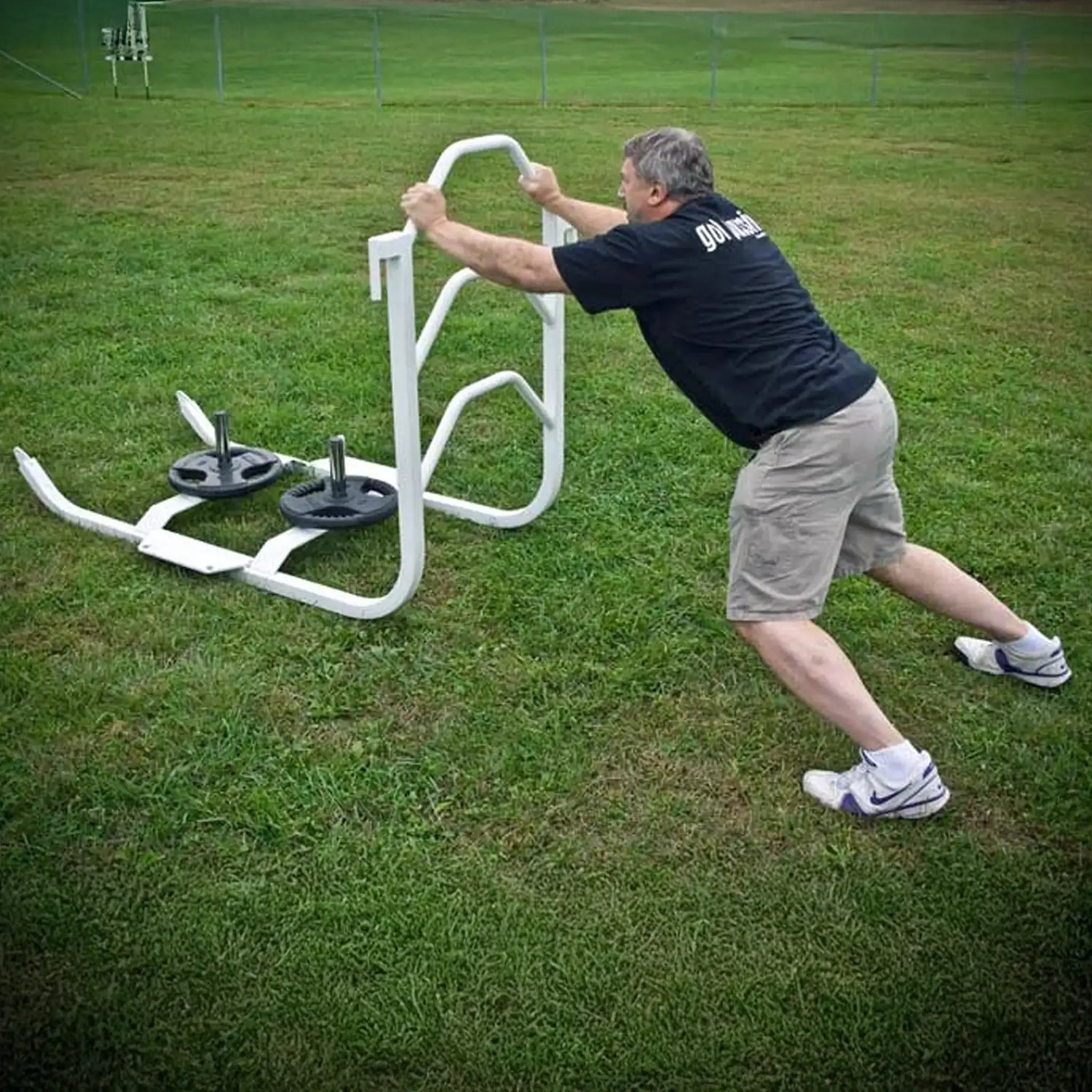 A man pushing a white metal push sled with black weight lifting plates on it on grass