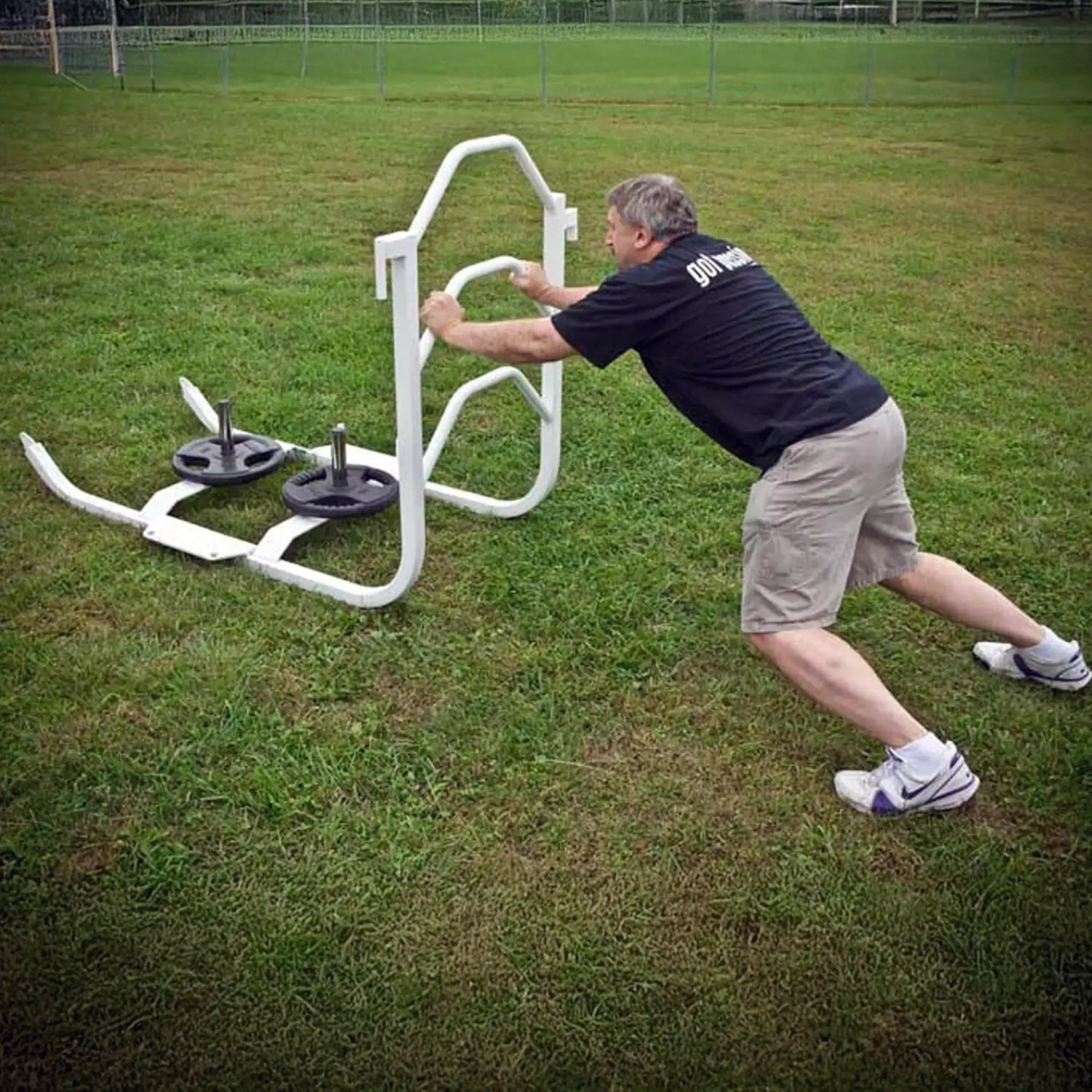 A man pushing a white metal push sled with black weight lifting plates on it on grass