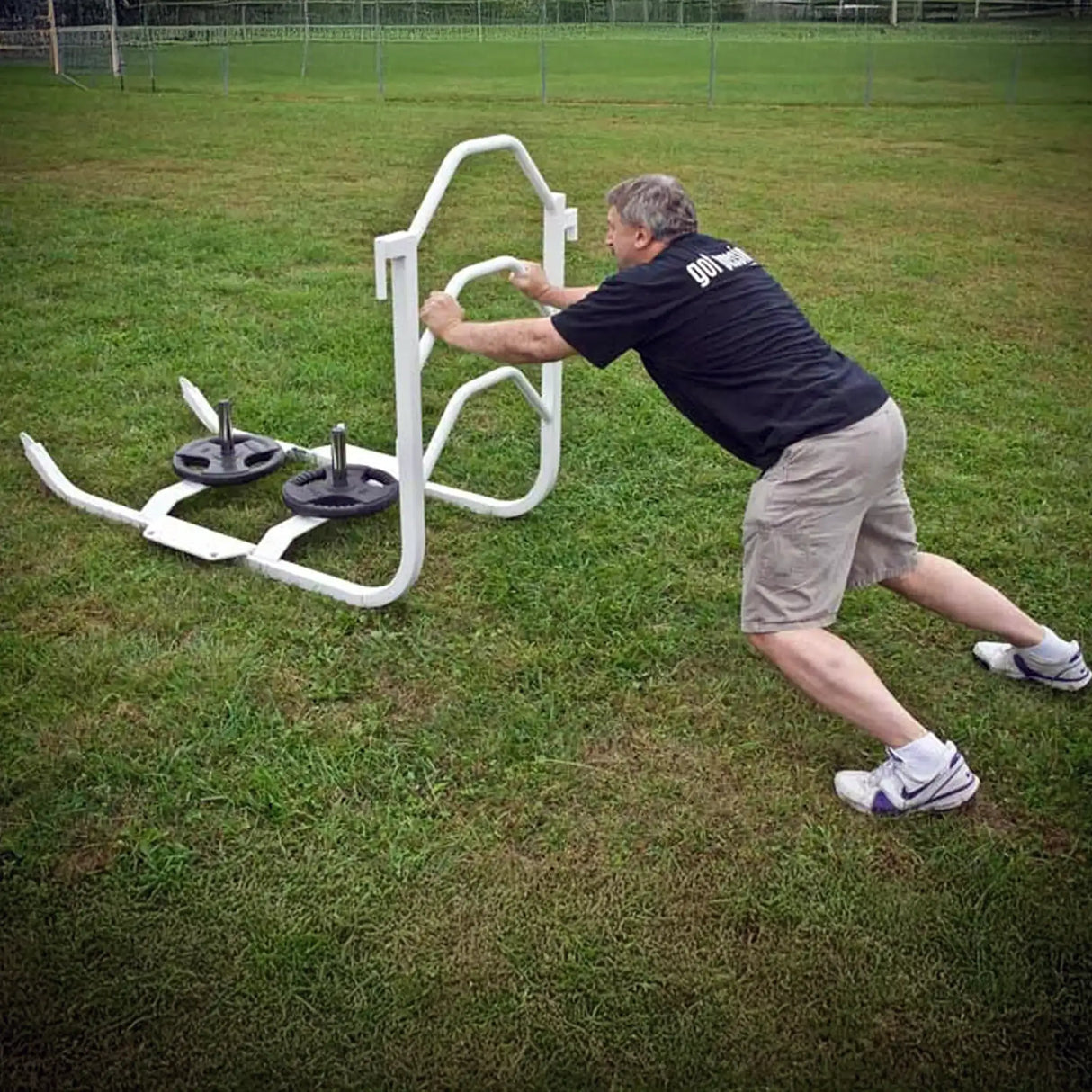A man pushing a white metal push sled with black weight lifting plates on it on grass