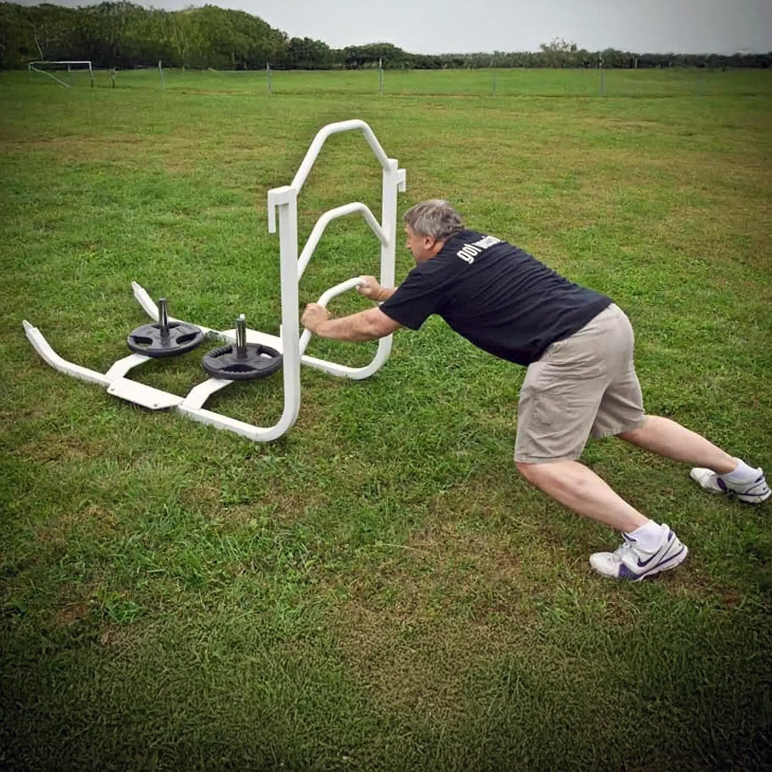 A man pushing a white metal push sled with black weight lifting plates on it on grass
