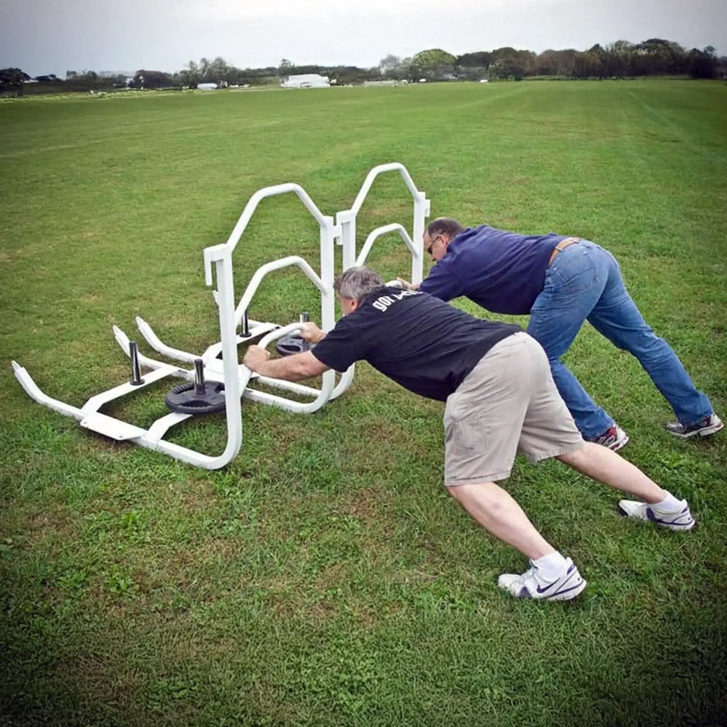 Two men pushing white metal push sleds with black weight lifting plates on them on grass