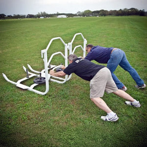 Two men pushing white metal push sleds with black weight lifting plates on them on grass