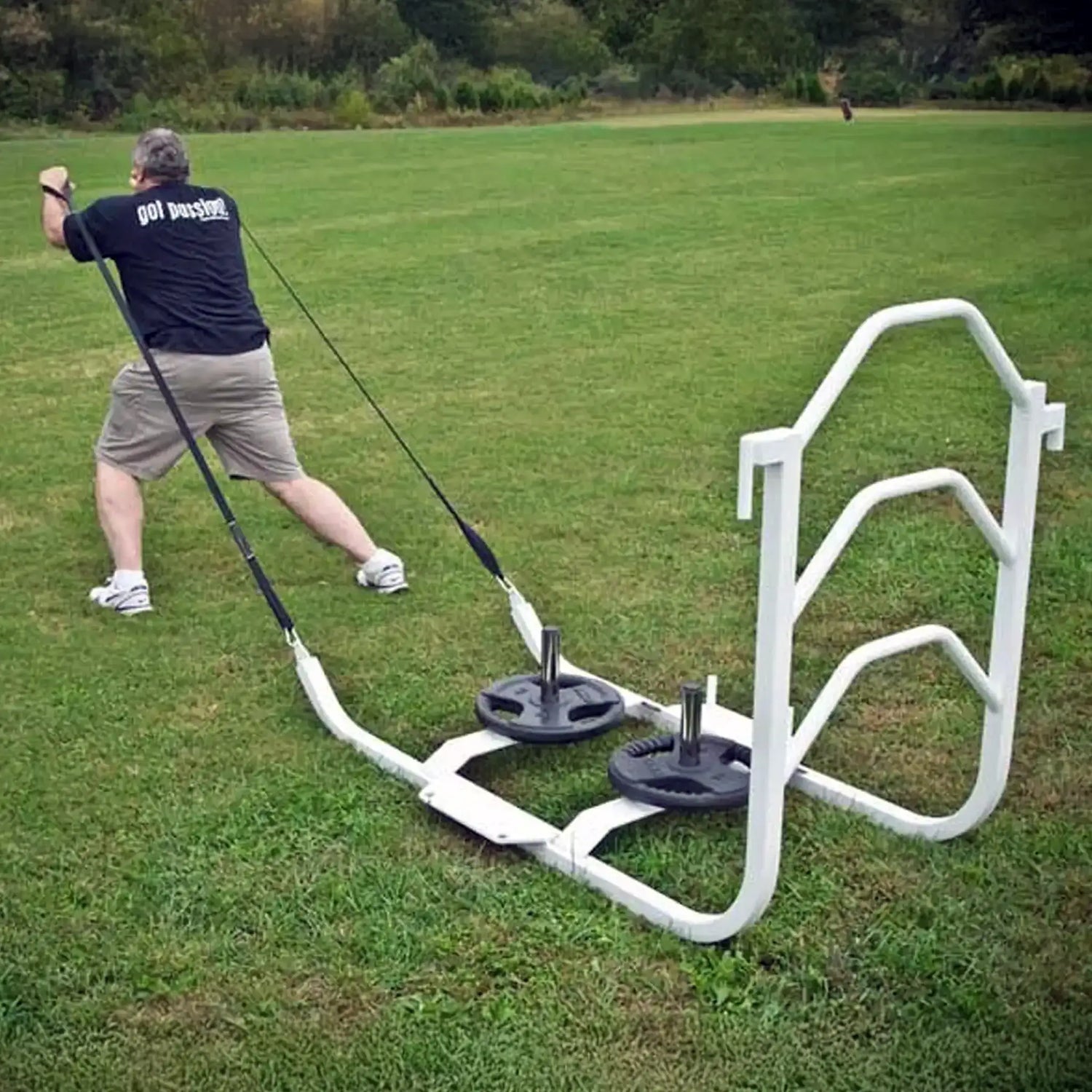 A man pulling a white metal sled on grass by two black straps