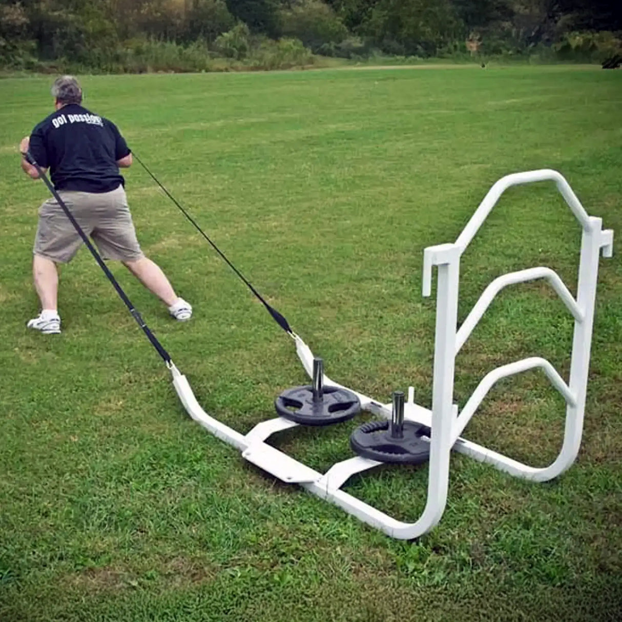 A man pulling a white metal sled on grass by two black straps