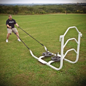 A man pulling a white metal sled on grass by two black straps