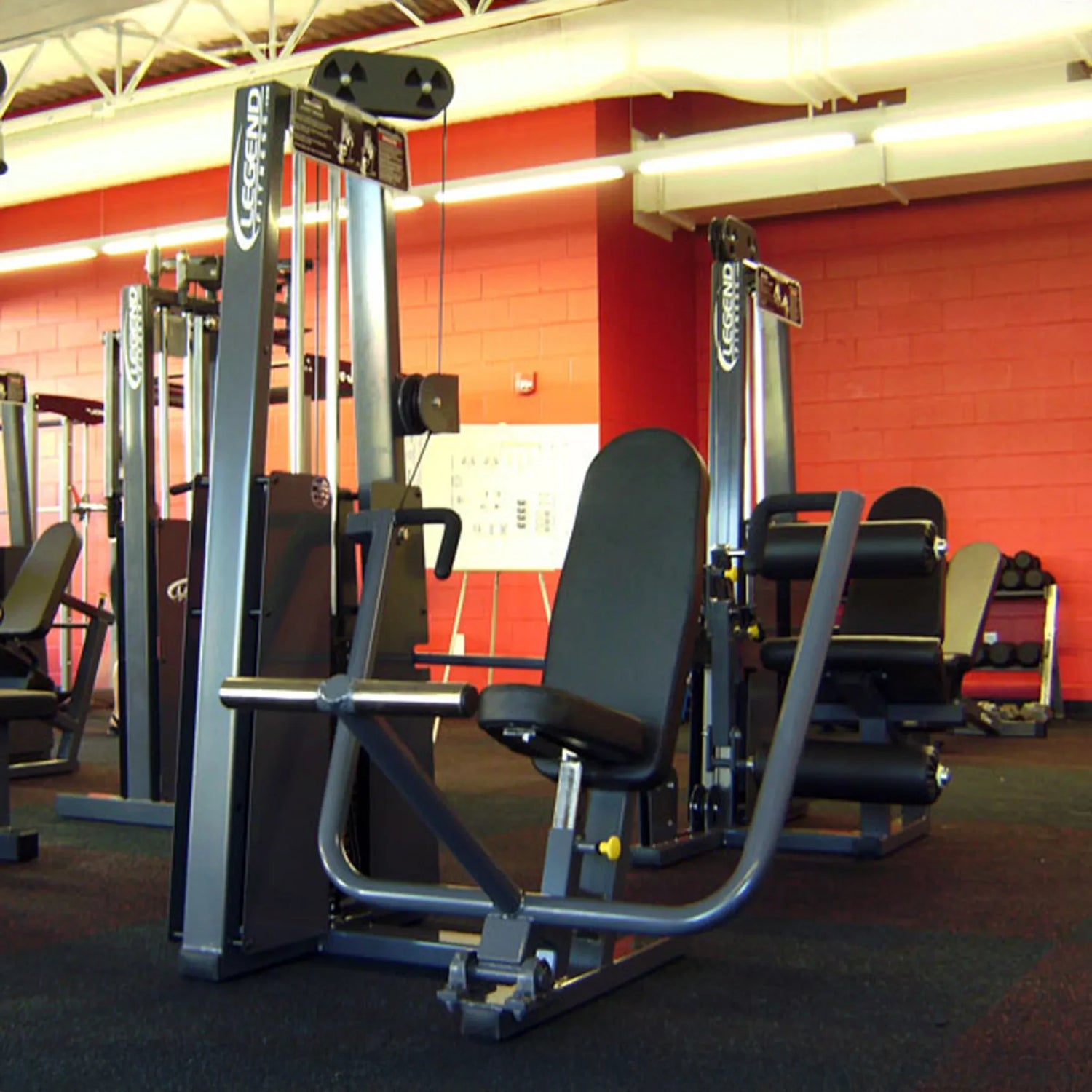 The Legend Fitness 900 Chest Press Machine by Legend Fitness features black padding and a metal frame, set on dark carpet in a brightly lit gym with red walls and other equipment in the background.