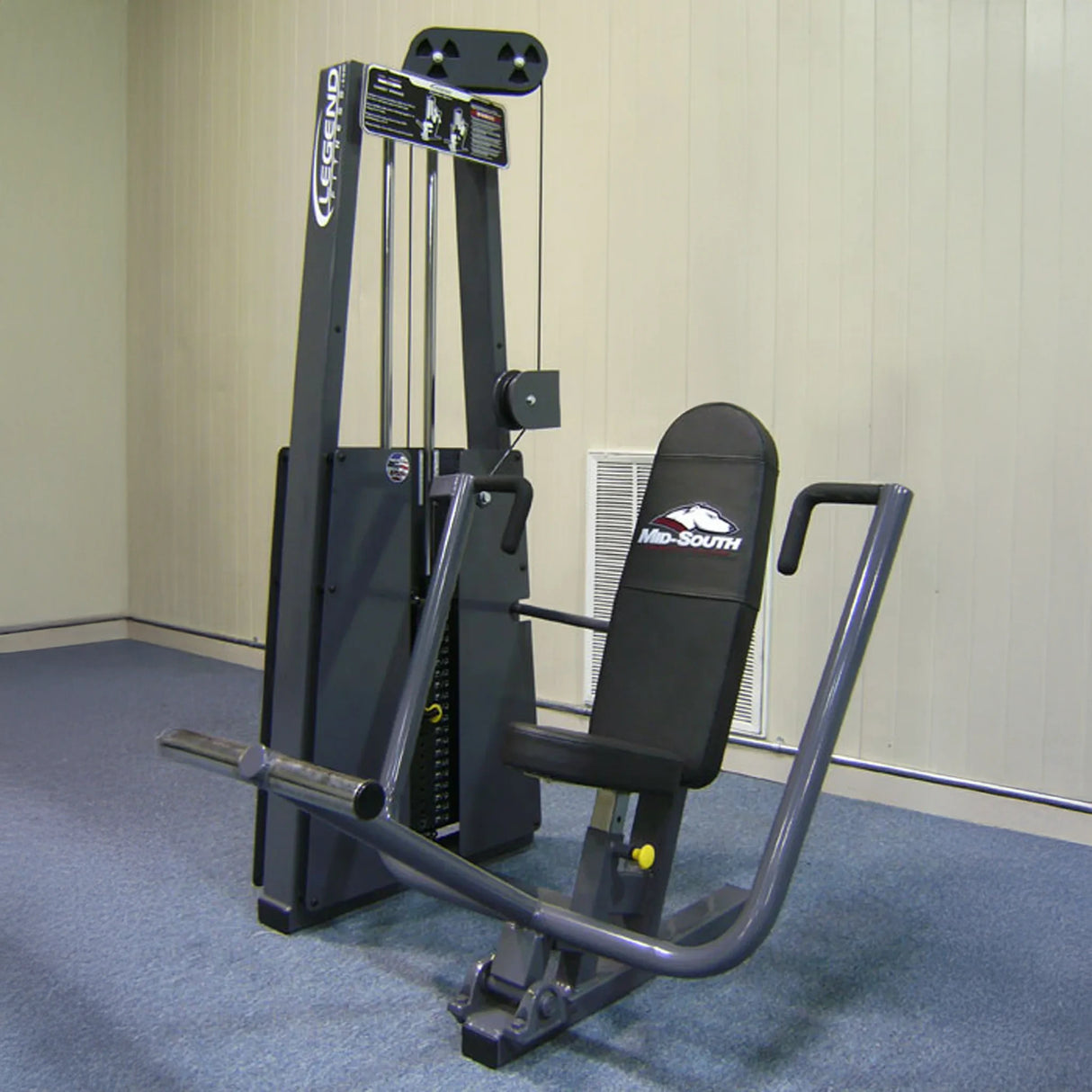 The Legend Fitness 900 Chest Press Machine by Legend Fitness features black padding, a gray metal frame, and adjustable weights and handlebars for strength training. It is placed on blue carpet in a room with beige walls.