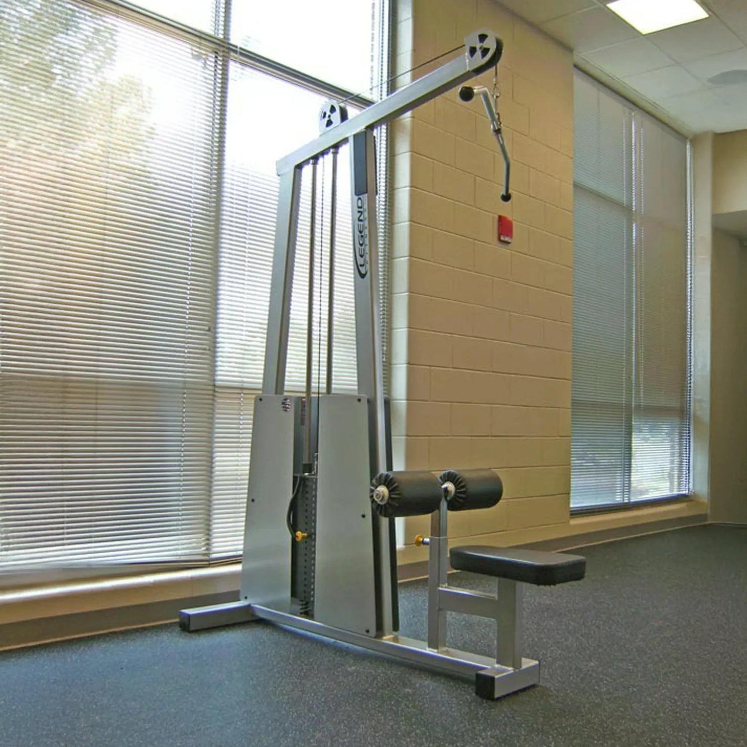 A Legend Fitness 905 Lat Pulldown Machine by Legend Fitness stands near a beige wall in a gym with large windows, blinds, black rubber flooring, and abundant natural light.