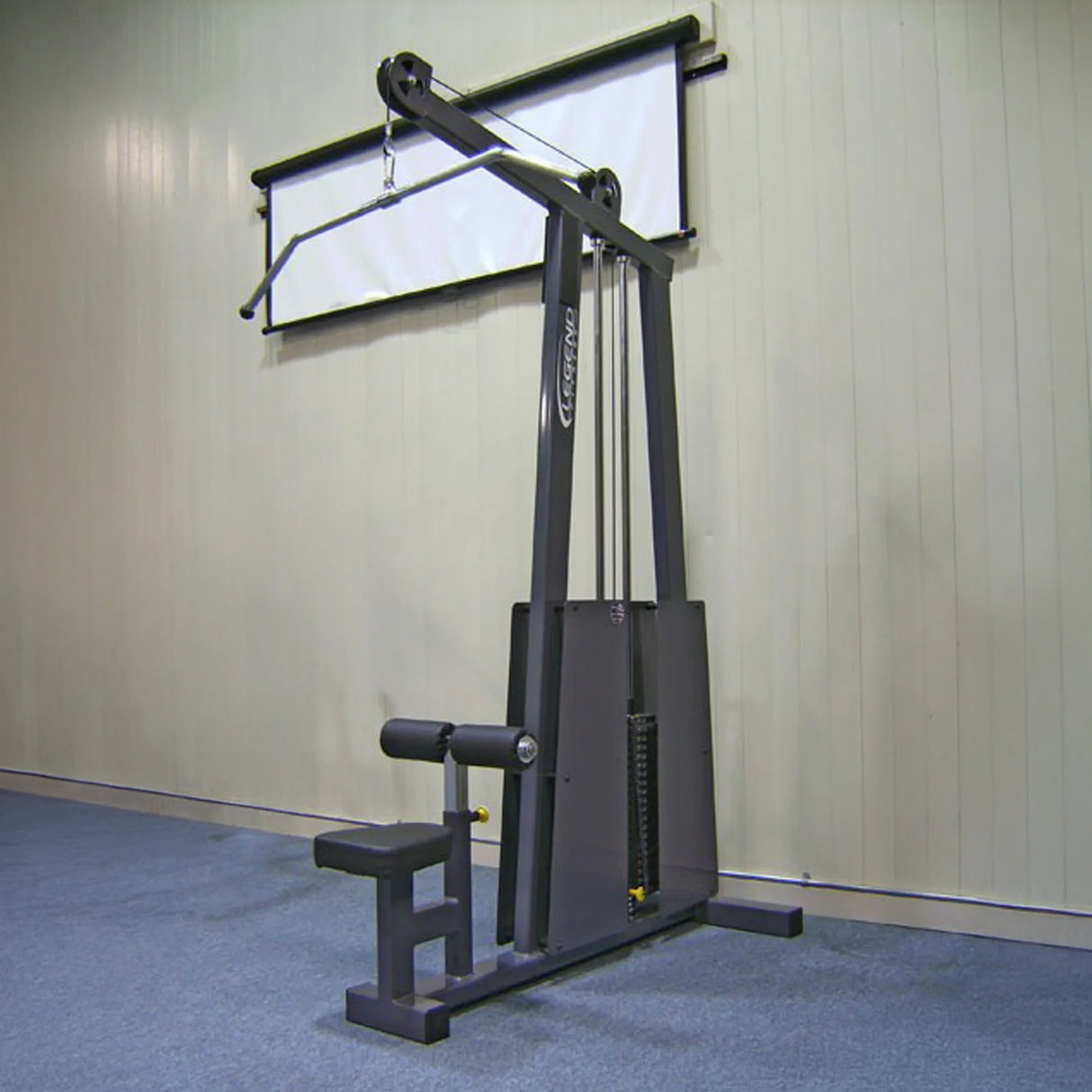 The Legend Fitness 905 Lat Pulldown Machine, featuring a padded seat and adjustable weights, is set against a light-colored gym wall with a white projection screen mounted above it.