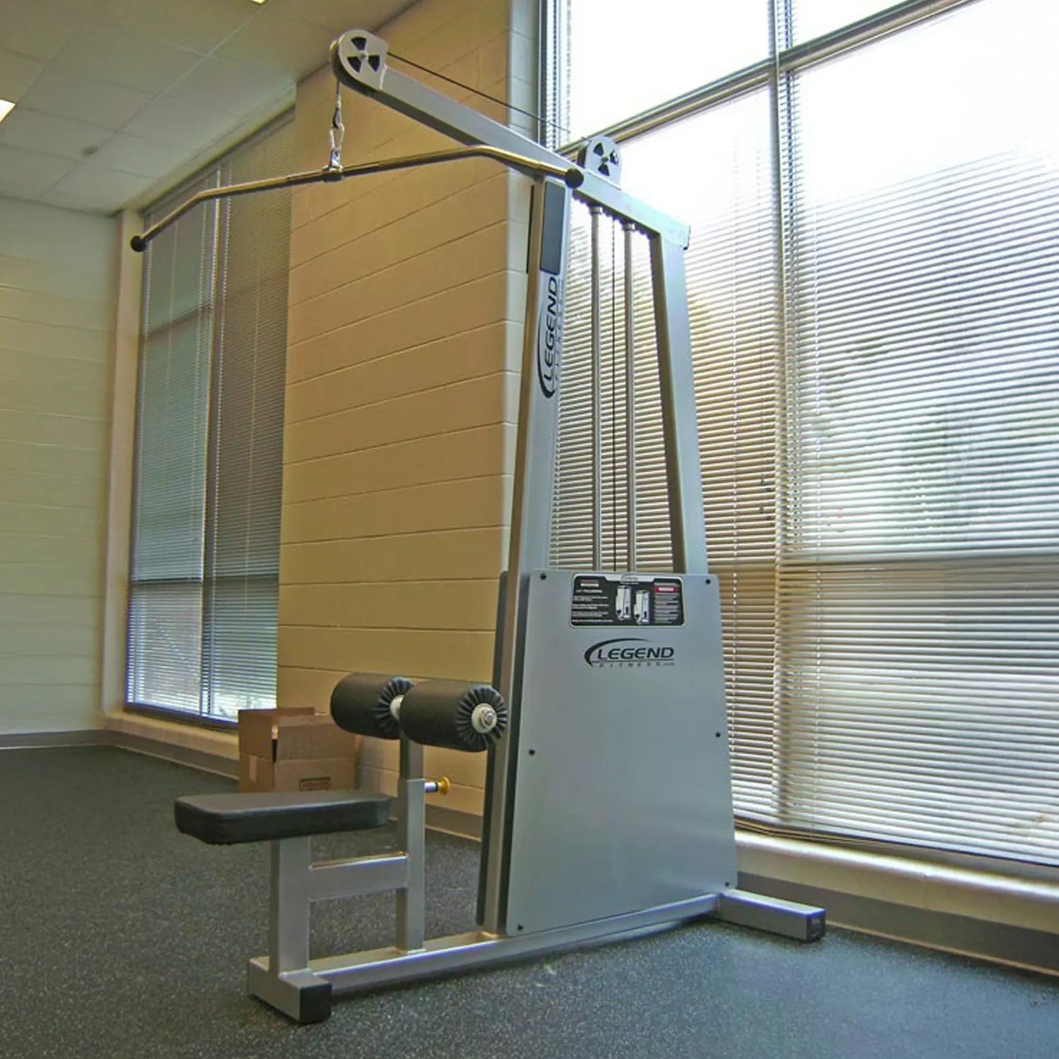 The Legend Fitness 905 Lat Pulldown Machine, featuring a silver finish, padded seat, and knee supports, is positioned in a gym by large windows with blinds that allow natural light to enter.