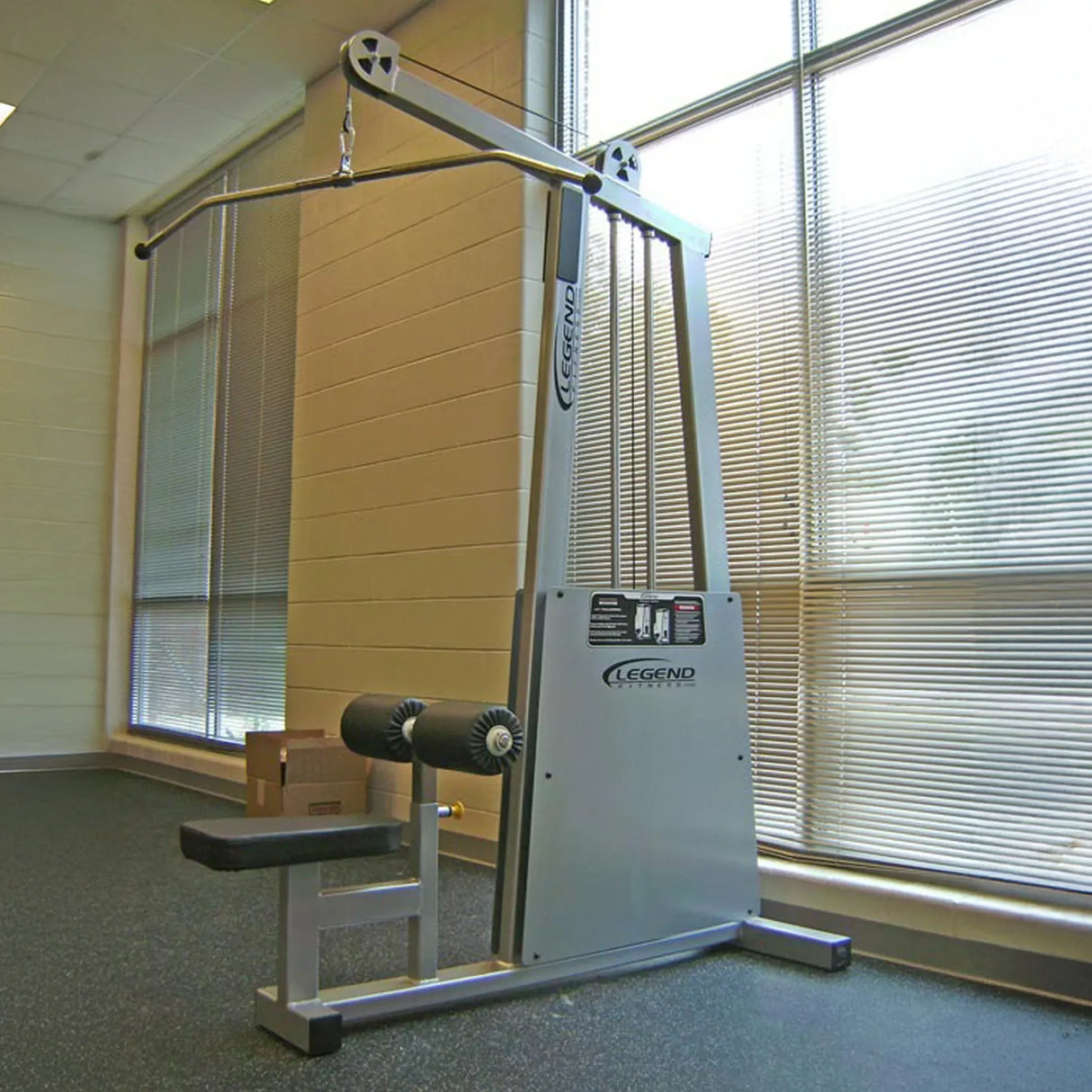 The Legend Fitness 905 Lat Pulldown Machine, featuring a silver finish, padded seat, and knee supports, is positioned in a gym by large windows with blinds that allow natural light to enter.