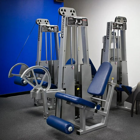 Two Legend Fitness 911 Leg Extension Machines by Legend Fitness, in blue and silver with padded seats and weights, are set up in a gym room with blue and white walls, gray carpet, and chairs partly visible on the right.