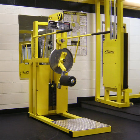 A Legend Fitness 916 Total Hip Combo Machine by Legend Fitness with weight plates stands in a gym, set against a white and black wall with mirrors and a shoulder workout poster in the background.