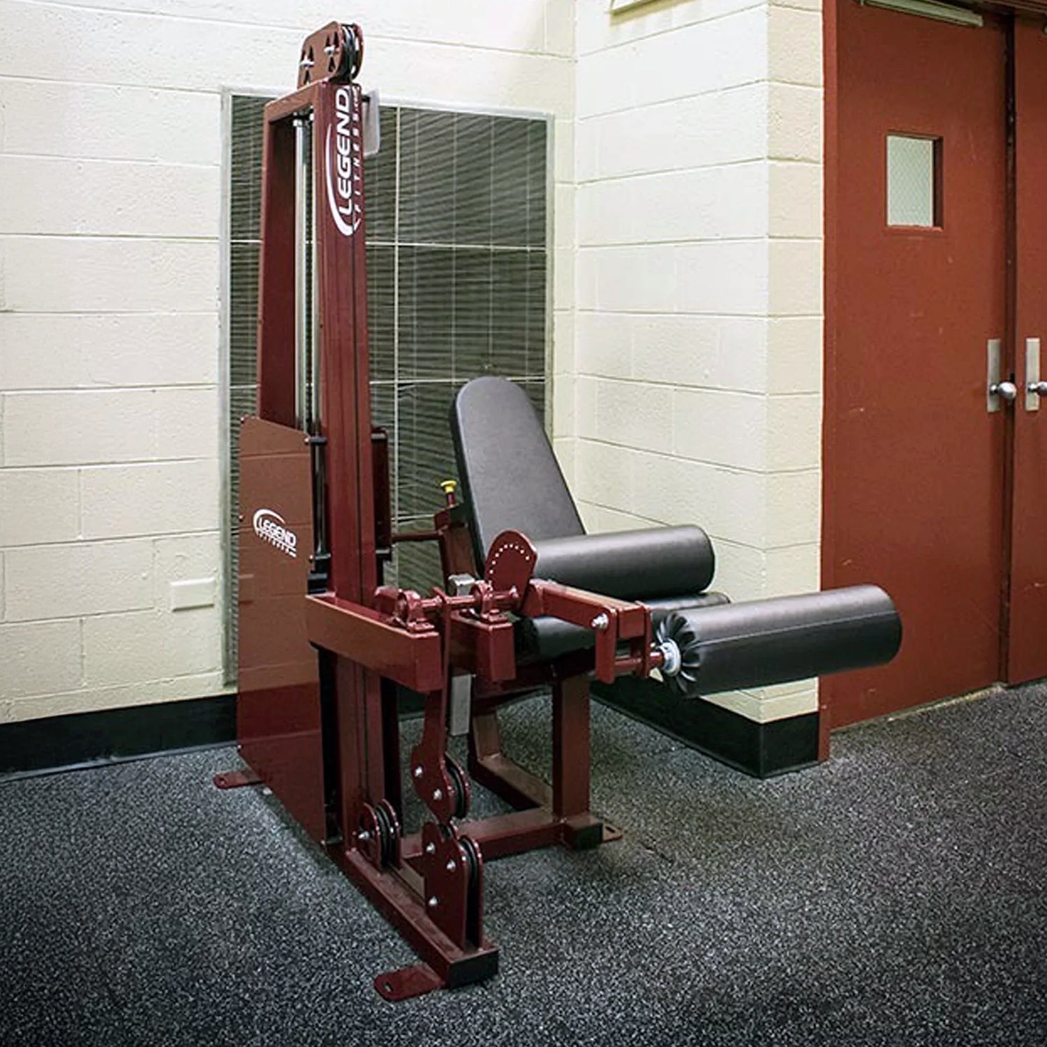 A Legend Fitness 918 Leg Extension / Leg Curl Combo Machine by Legend Fitness is set in a gym with beige walls, featuring a maroon padded bench, padded leg rollers for support, and a door visible in the background.