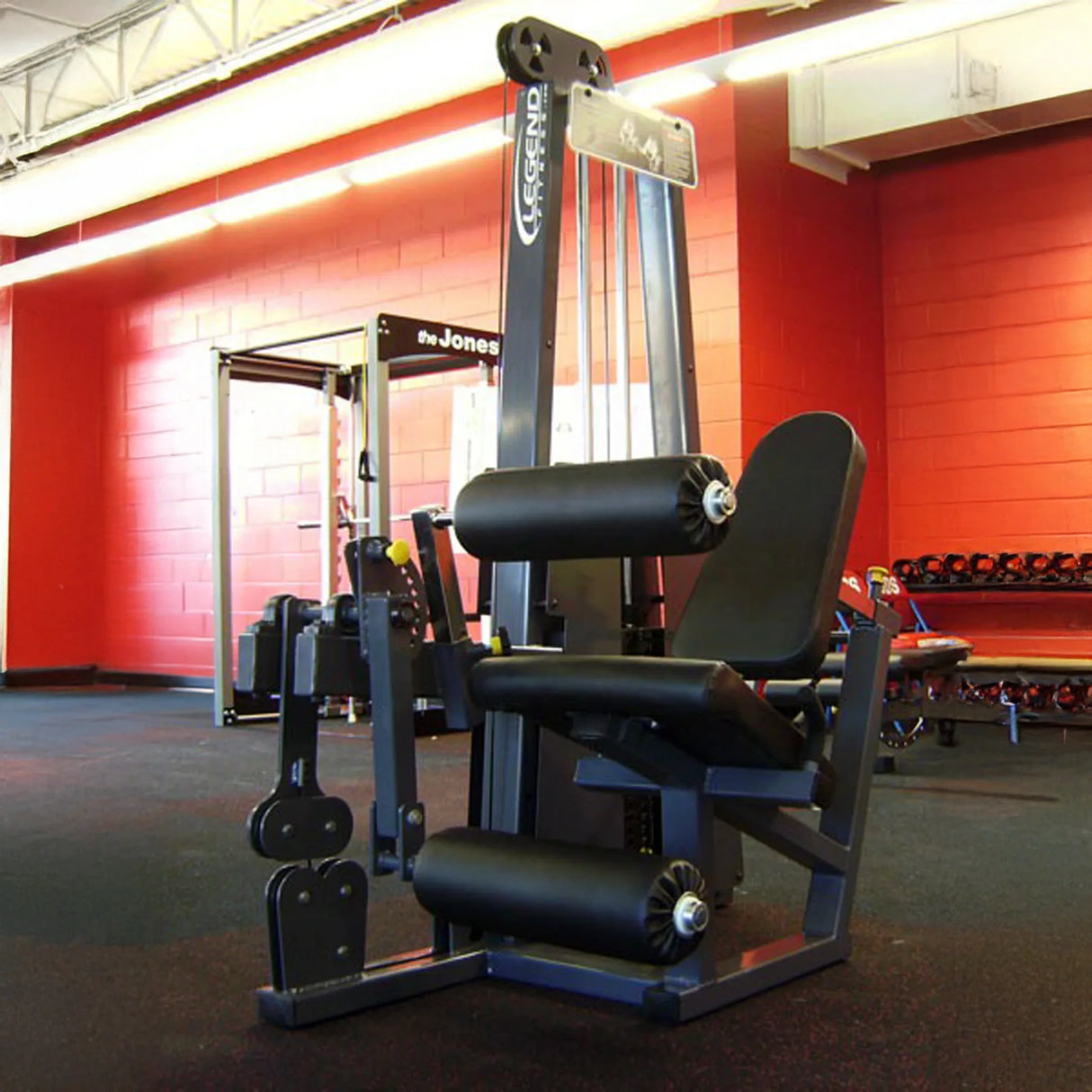 The Legend Fitness 918 Leg Extension / Leg Curl Combo Machine by Legend Fitness sits on a gym floor with red and tan walls, surrounded by assorted fitness equipment and free weights.