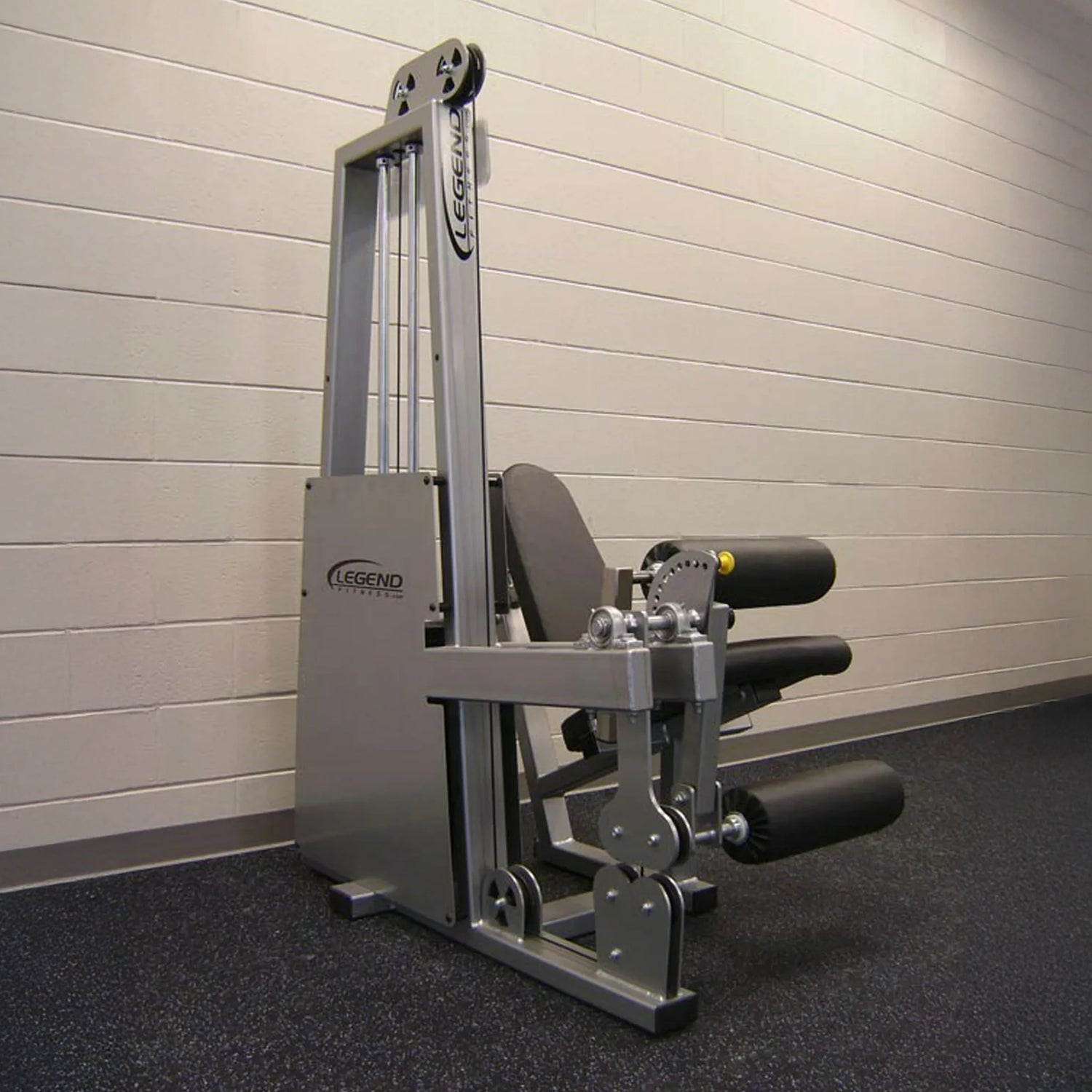 The Legend Fitness 918 Leg Extension / Leg Curl Combo Machine by Legend Fitness, featuring padded black rollers and a silver frame, is set against a beige brick wall on a dark speckled gym floor.