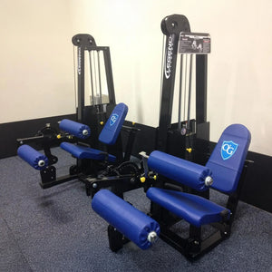 Two Legend Fitness 918 Leg Extension/Leg Curl Combo Machines in blue and black, featuring padded seats and rollers, are placed side by side on grey carpet against white walls.