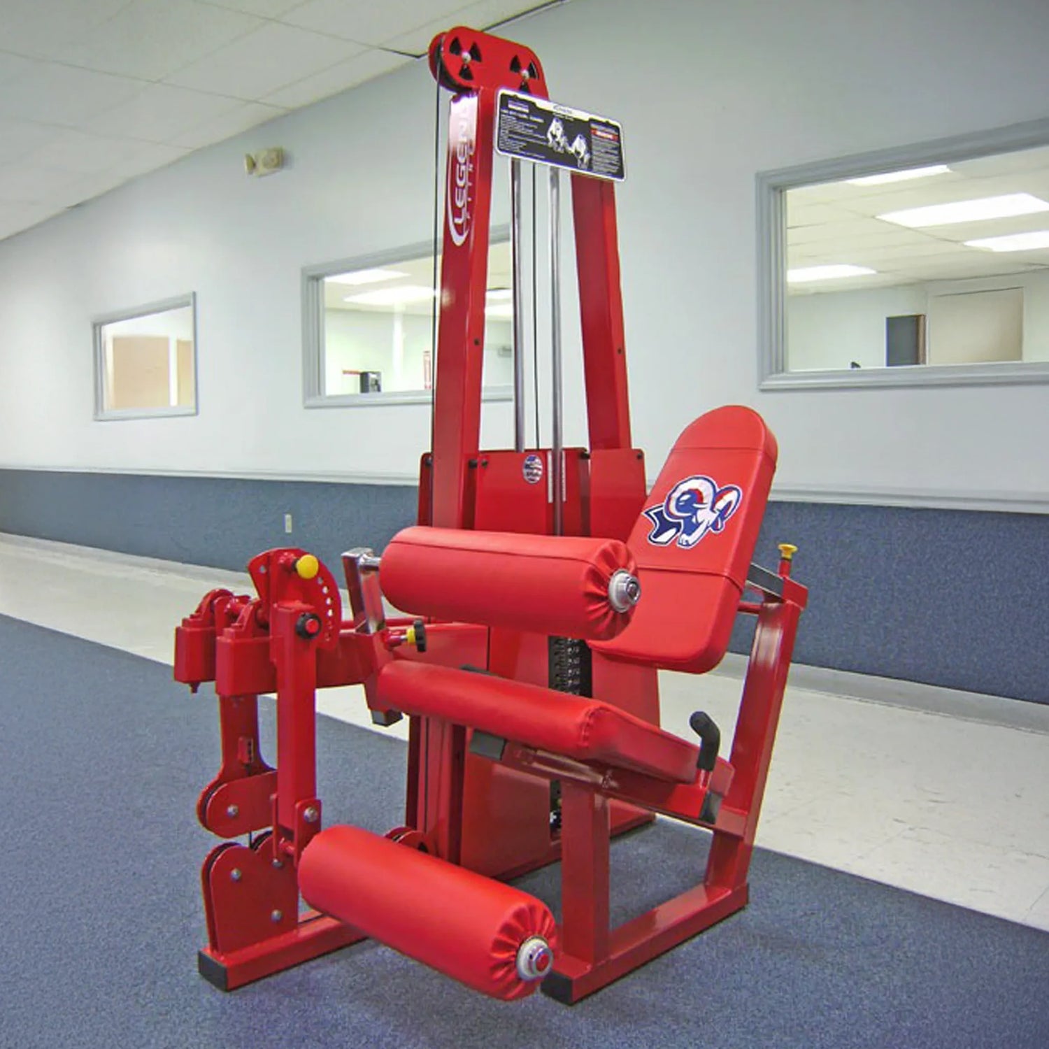 A Legend Fitness 918 Leg Extension / Leg Curl Combo Machine with padded rollers and a logo on the backrest stands in a gym with blue carpet, white walls, and windows into adjacent rooms.