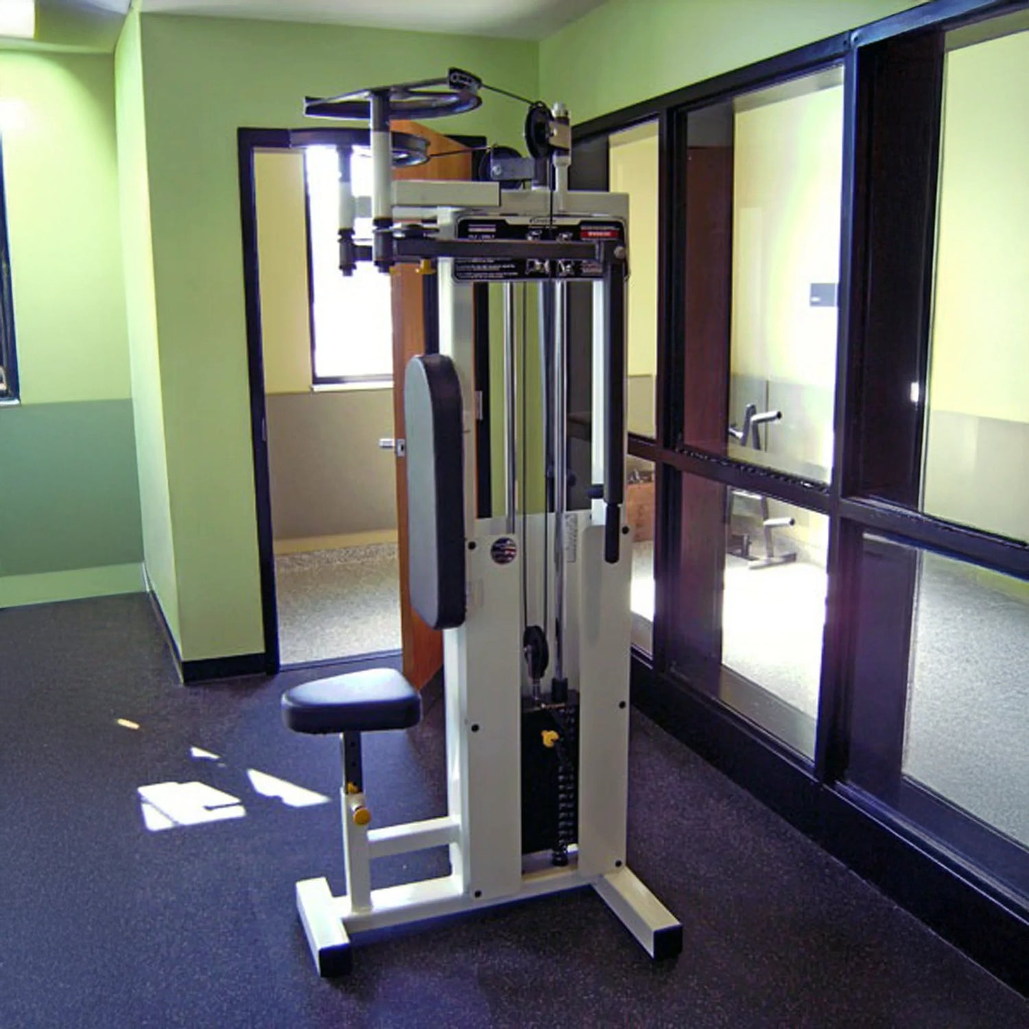 A Legend Fitness 942 Rear Delt / Pec Combo Machine with a padded seat and backrest stands in a small, brightly lit gym room featuring green walls, black rubber flooring, and large windows.