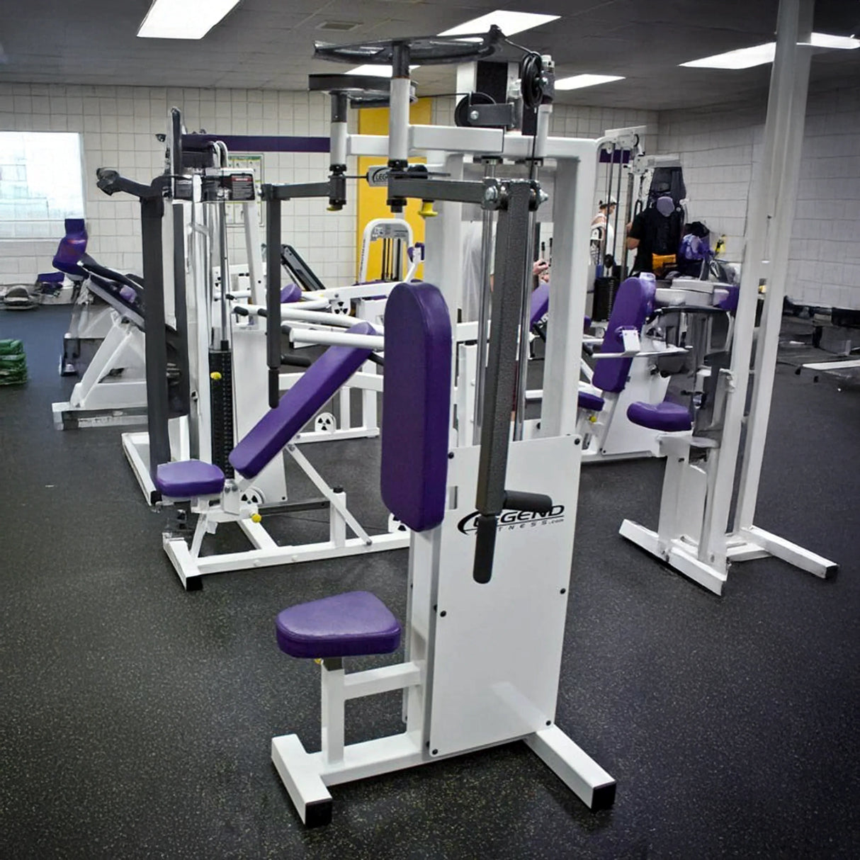 A brightly lit gym features the Legend Fitness 942 Rear Delt / Pec Combo Machine, all white with purple padding, set on a rubber floor among other strength training equipment.