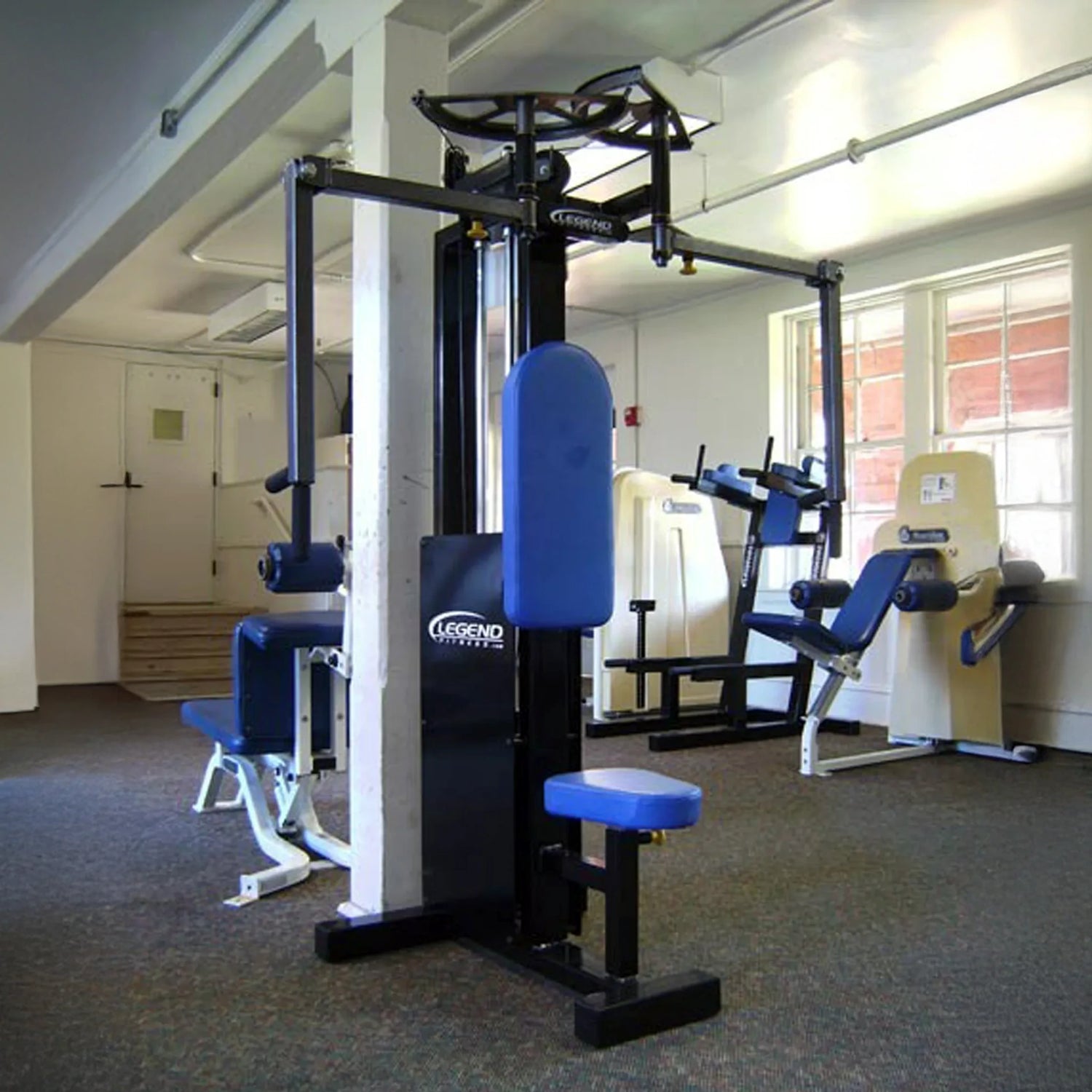 A gym room with various exercise machines, featuring a blue and black Legend Fitness 942 Rear Delt / Pec Combo Machine in the foreground and other workout equipment near the windows in the background.