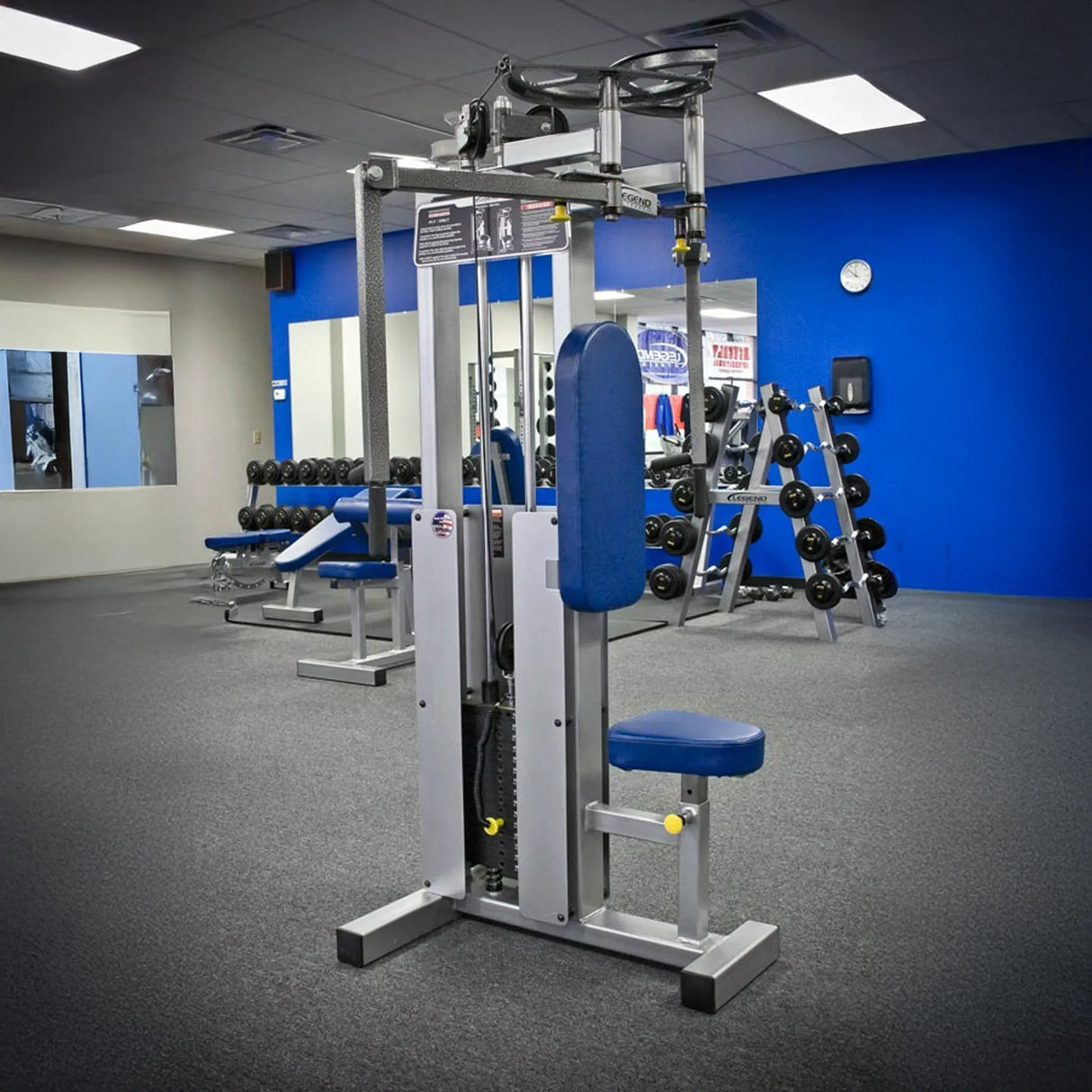 A gym with gray carpet, blue and white walls, free weights, and mirrors features a Legend Fitness 942 Rear Delt / Pec Combo Machine with blue padding as the centerpiece among various weight machines.