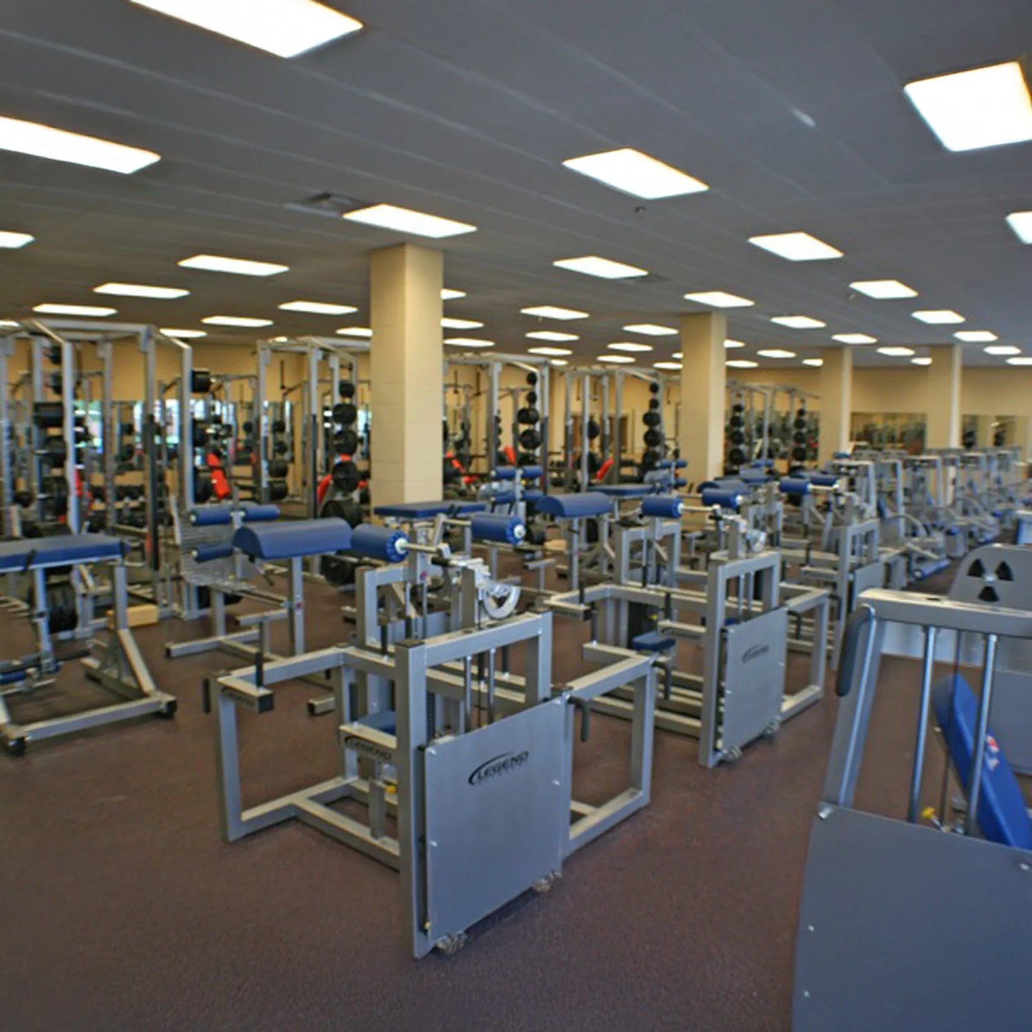 A spacious gym features multiple Legend Fitness 943 Neck Machine machines and benches in rows under bright overhead lights, with mirrors lining the back wall.