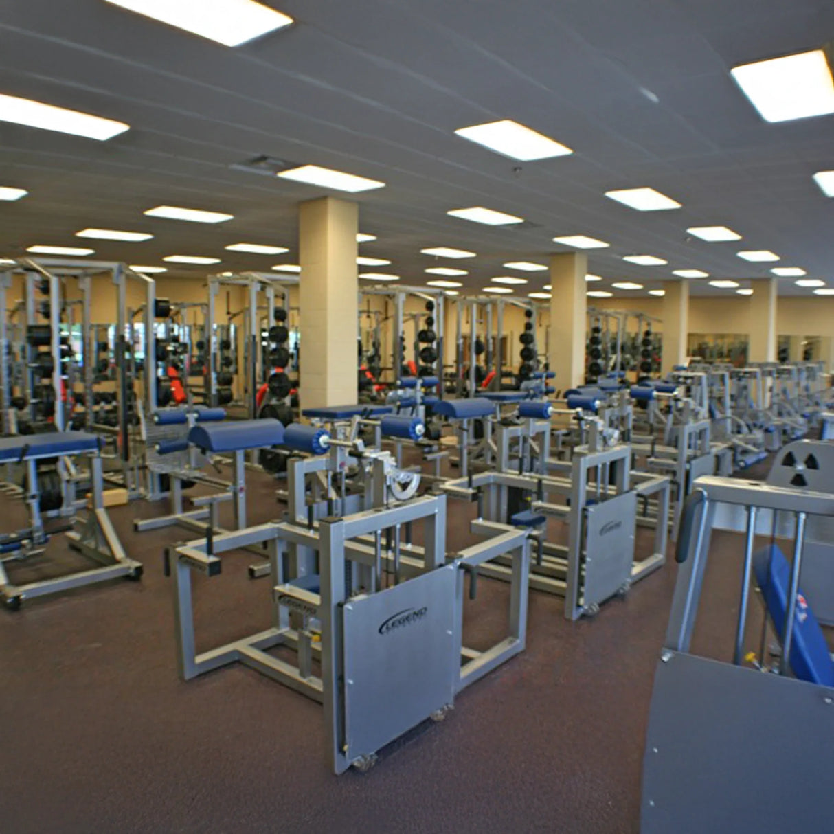 A spacious gym features multiple Legend Fitness 943 Neck Machine machines and benches in rows under bright overhead lights, with mirrors lining the back wall.