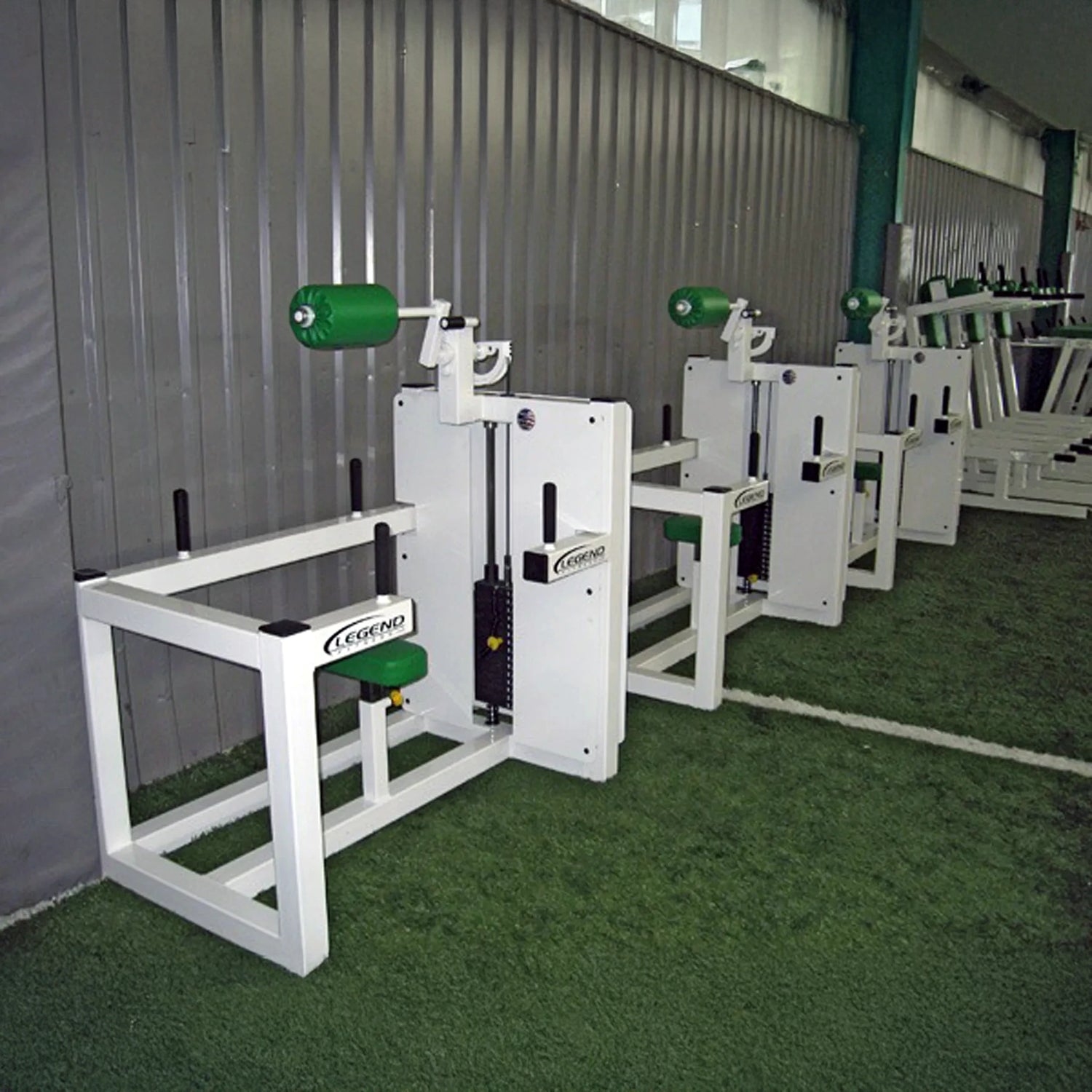 Four Legend Fitness 943 Neck Machines with green padding are lined up on artificial turf beside a corrugated metal wall in an indoor gym.