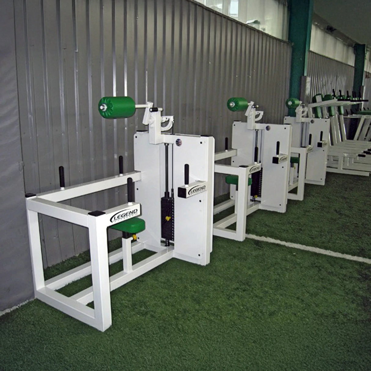 Four Legend Fitness 943 Neck Machines with green padding are lined up on artificial turf beside a corrugated metal wall in an indoor gym.