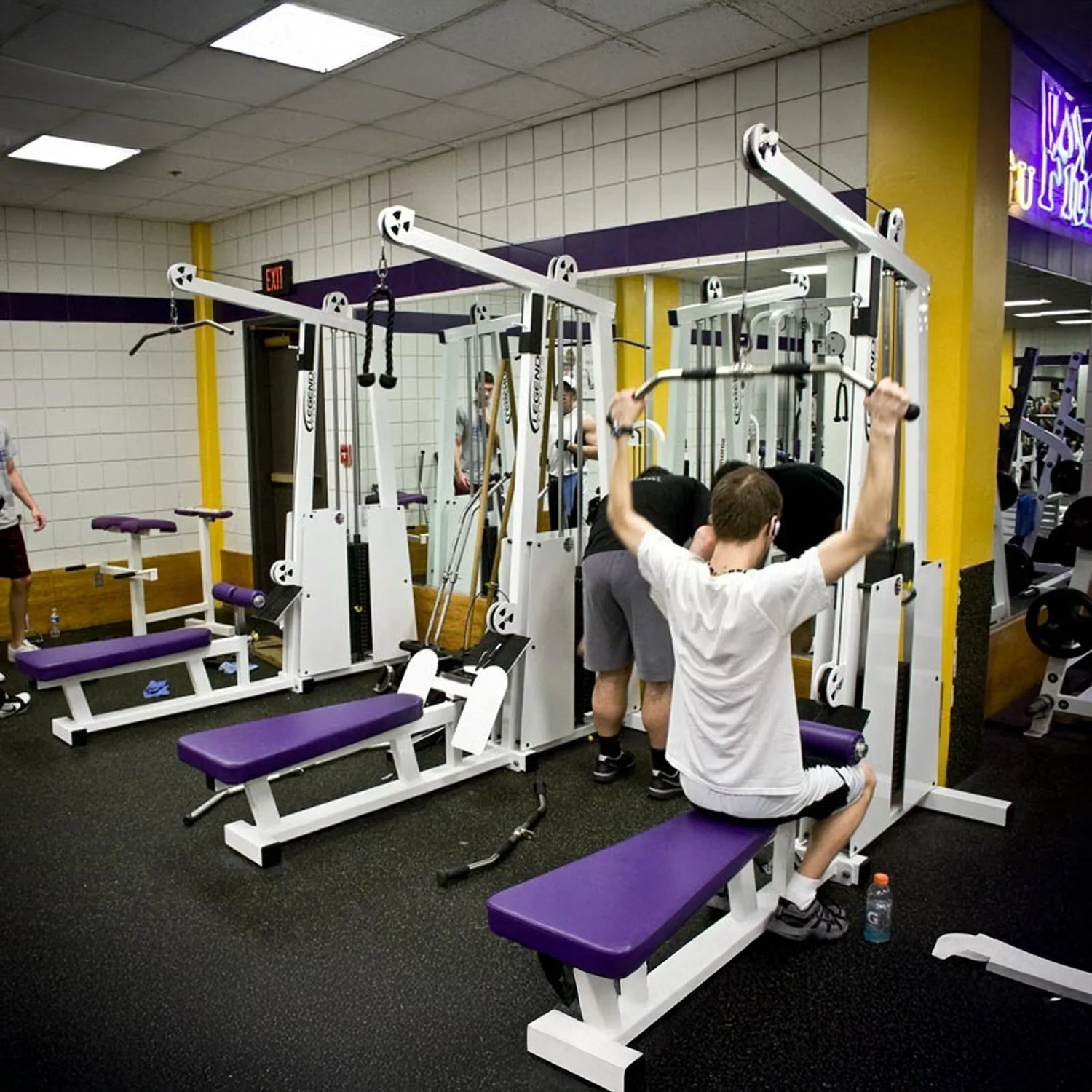 A person uses the Legend Fitness 945 Lat Pull / Low Row Combo Machine in a gym with purple benches, white exercise equipment, mirrored and white tiled walls, while others are visible in the background.